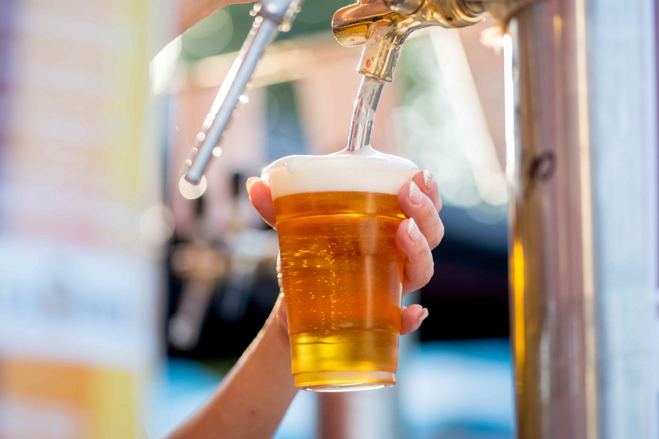 Hand pouring a draft beer from a tap into a plastic cup, with foam forming on top at a bar or event beverage station.