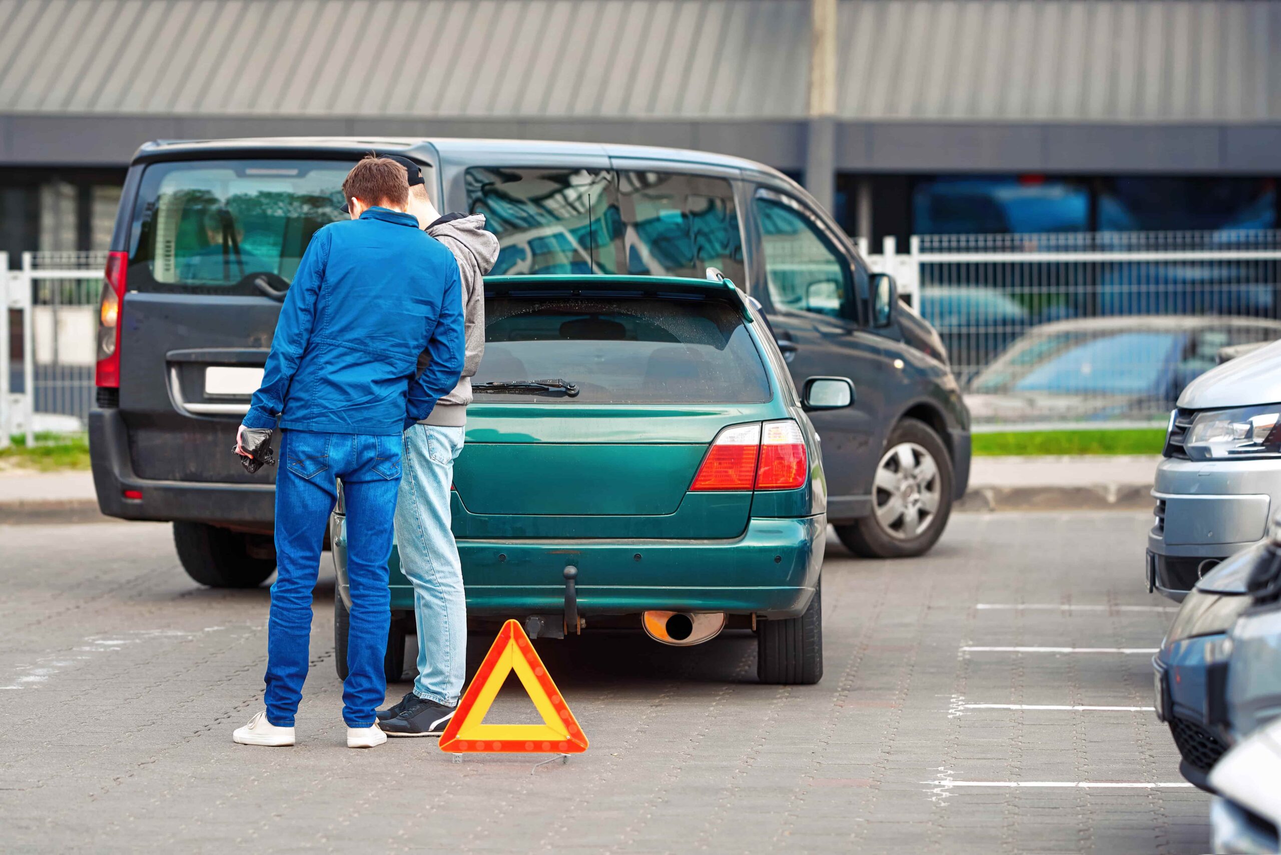 Two men inspecting a broken-down car with a warning triangle placed behind the vehicle in a parking lot after a minor car breakdown.