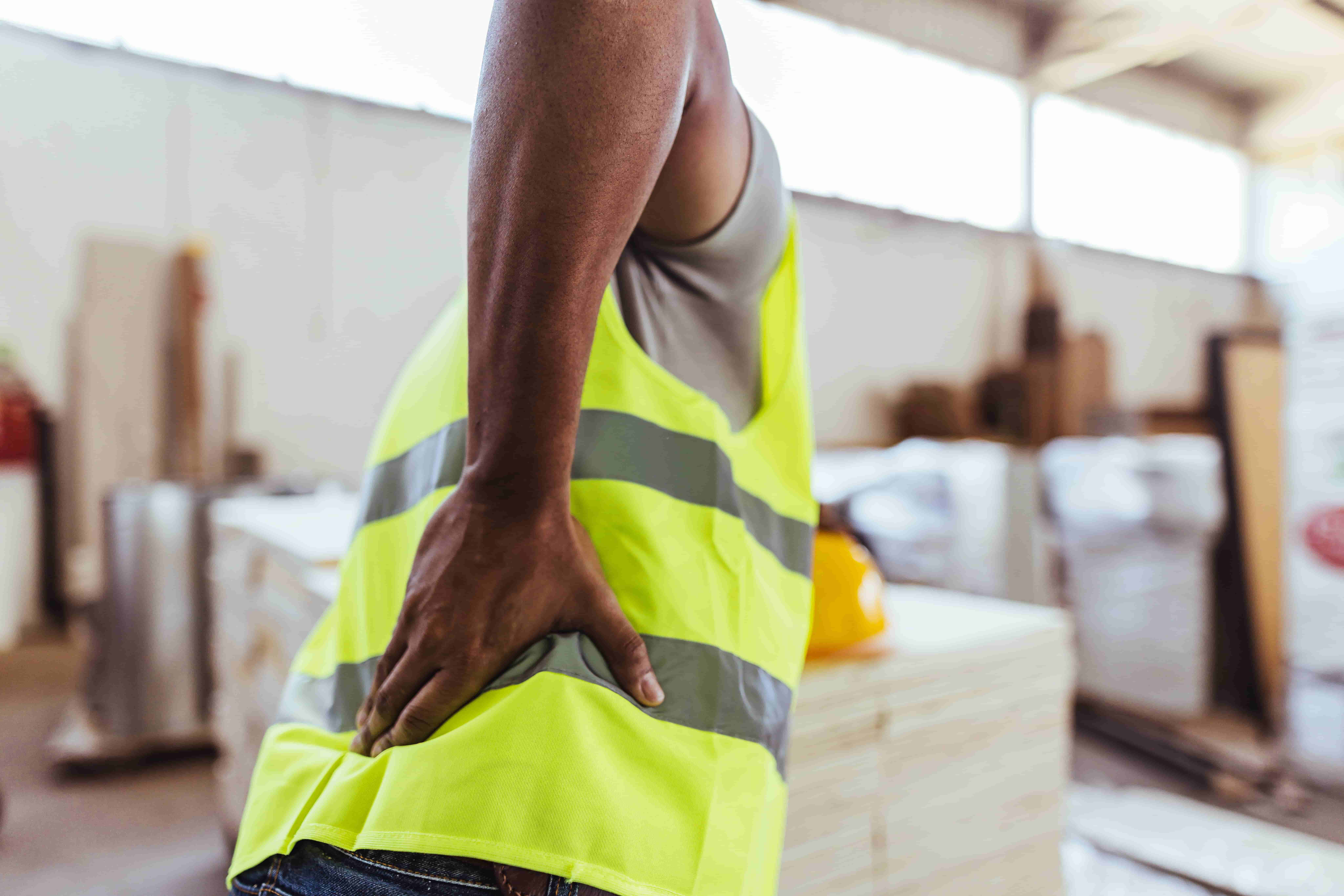 Construction worker experiencing lower back pain while wearing a safety vest at an industrial job site.